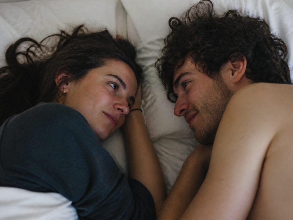 Couple allongé face à face sur lit blanc, lumière tamisée ambiante